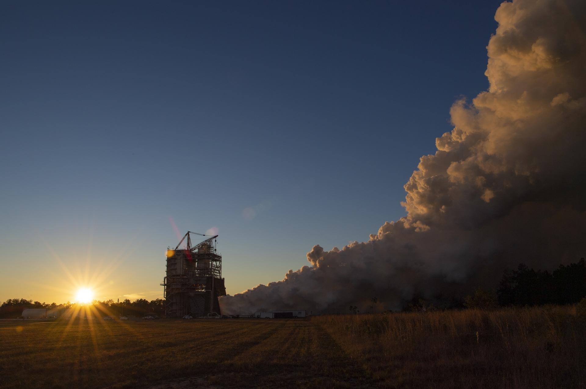 A rocket engine test stand firing at dusk, exhaust plume lit by the sun